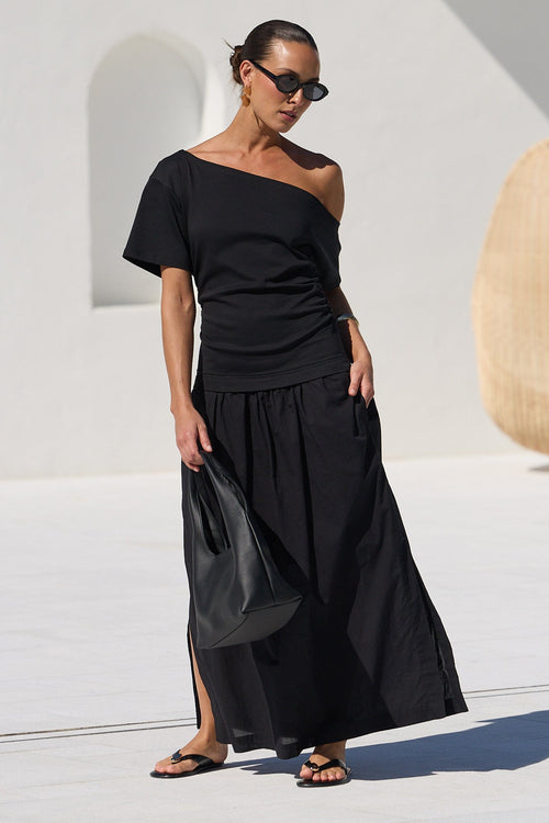 A woman wearing a black off-the-shoulder top and matching black wide-leg skirt stands on a white patio poolside, holding a black handbag. The background features a modern white architectural structure with a hanging woven chair and a tiled swimming pool.