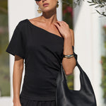 A woman wearing a black asymmetrical short-sleeve top and black wide-leg pants, carrying a large black leather hobo shoulder bag. She is wearing black sunglasses and gold earrings, standing outdoors in front of a white building with olive tree branches overhead.