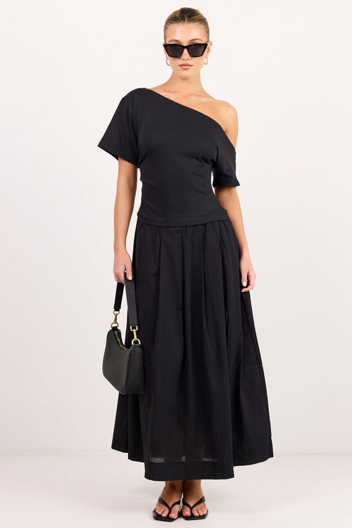 A model wearing a black one-shoulder midi dress with a peplum waist, holding a black handbag, set against a plain white studio background.