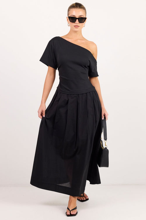 A woman modeling a black one-shoulder midi dress with short sleeves, a fitted bodice, and a flowing skirt, styled with black sunglasses, black sandals, and a small black handbag against a plain white studio background.