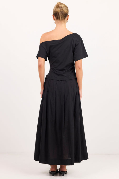 A woman viewed from the back wearing a black off-the-shoulder midi dress with short sleeves and a pleated skirt, standing against a plain white studio background.