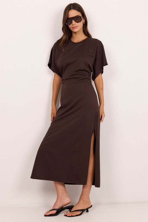A woman modeling a dark brown short-sleeve midi dress with a high side slit, styled with black sunglasses and black heeled sandals against a plain white studio background.