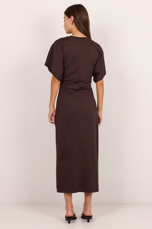 A woman viewed from the back wearing a dark brown short-sleeve midi dress with a gathered waist, paired with black mule heels, against a plain off-white studio background.