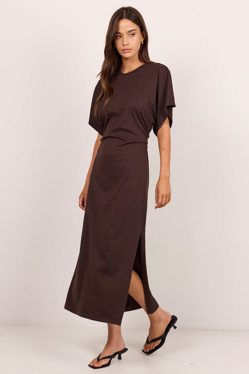 A woman modeling a dark brown short-sleeve maxi dress with a high side slit, paired with black heeled sandals, against a plain off-white studio background.