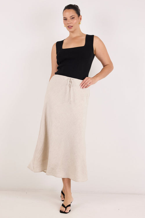 A woman modeling a black sleeveless top and a long, flowing cream-colored linen skirt with a drawstring waist, paired with black strappy heels, against a plain white studio background.