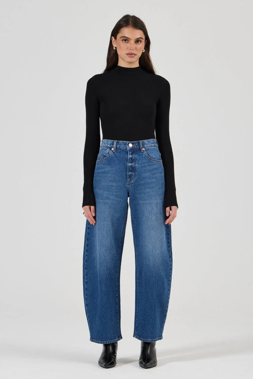 A model stands against a light gray studio background wearing a black long-sleeve mock-neck top, high-waisted wide-leg blue jeans, and black ankle boots.