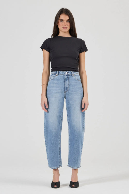 A woman modeling a pair of light-wash blue cropped jeans with a black short-sleeve t-shirt and black flat shoes, standing against a light gray studio background.