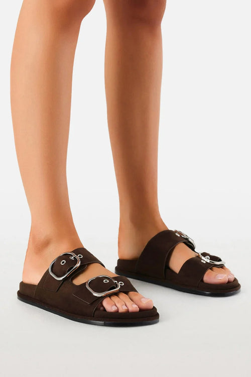 A person wearing dark brown double-strap slide sandals with large silver-tone buckle details, shown from the calves down against a clean light gray studio background.