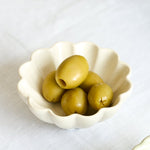 Two small scalloped ceramic bowls on a white fabric surface, with one bowl containing green olives and the other empty, styled with olive branches in the background.