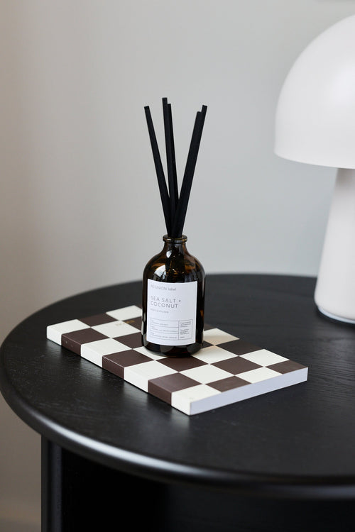 An amber glass reed diffuser bottle with black reeds stands on a black-and-white checkerboard tray on a round black table, with a white lamp and a white cup with gold trim in the background.