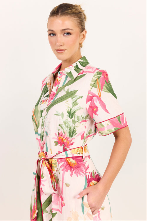 A woman modeling a short-sleeve collared dress with a pink, green, and orange tropical floral print on a white base, featuring a matching tie belt with a gold buckle.
