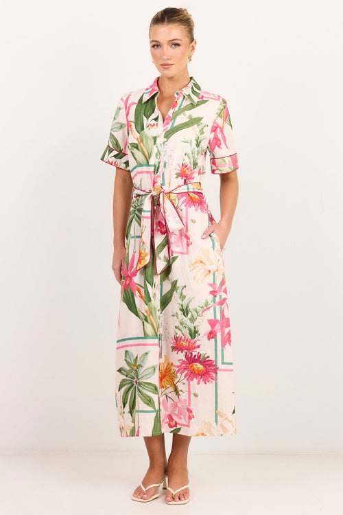A woman modeling a short-sleeve collared midi dress with a tropical floral print, featuring pink flowers, green leaves, and a cream background with a matching tie belt, set against a plain white studio backdrop.