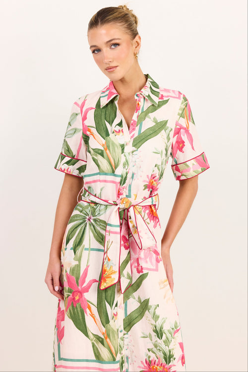 A woman modeling a short-sleeve collared dress with a tropical floral and leaf print on a light background, featuring a matching tie belt, against a plain light studio backdrop.