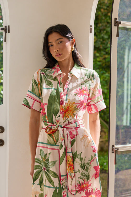 A woman wearing a short-sleeve collared shirt dress with a colorful tropical floral and leaf print, standing in front of white arched doorways with greenery visible outside.