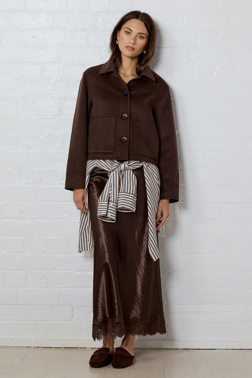 Woman wearing a brown wool jacket with a satin brown skirt leaning on a white brick wall background
