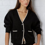 Woman wearing a black cardigan with white trim against a white brick wall.