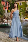 Woman in a blue dress standing by a waterfront with boats and greenery in the background