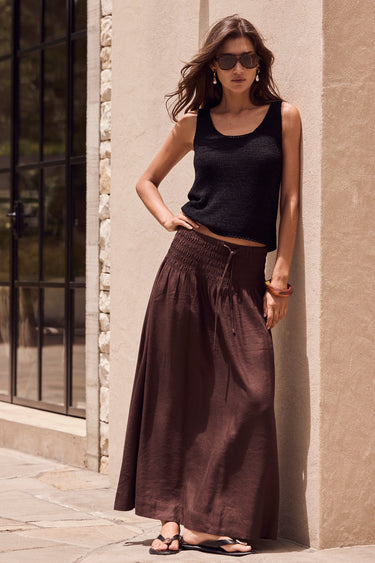Woman in black tank top and brown wide-leg pants standing against a beige wall.