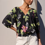 A woman models a black short-sleeve blouse with a green and pink tropical floral print, paired with light beige shorts, standing against a minimalist beige wall with a strong shadow.