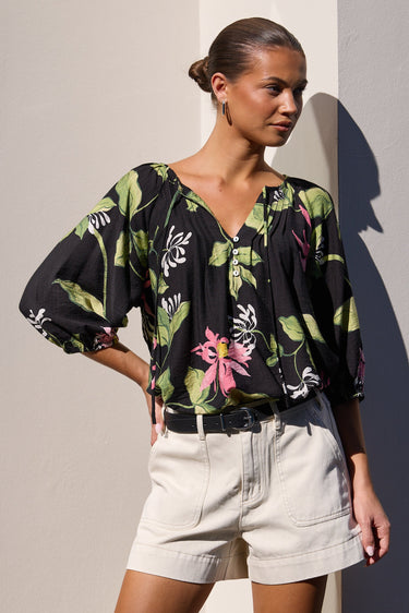 A woman models a black short-sleeve blouse with a green and pink tropical floral print, paired with light beige shorts, standing against a minimalist beige wall with a strong shadow.
