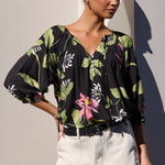 A woman models a black blouse with a green and pink tropical floral print, paired with light beige shorts, standing against a minimalist beige wall with a strong diagonal shadow.