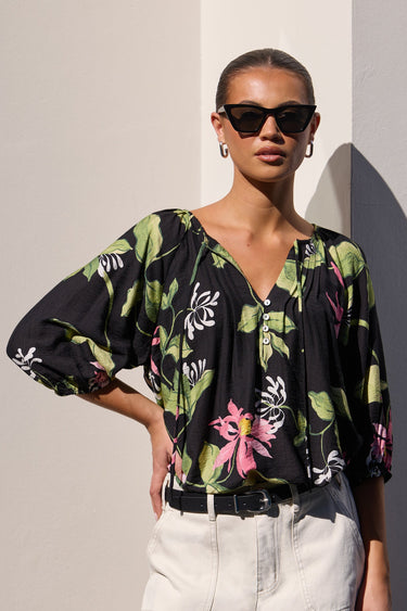 A woman modeling a black floral blouse with green and pink botanical print, paired with white shorts, standing against a light wall with strong sunlight and shadows.