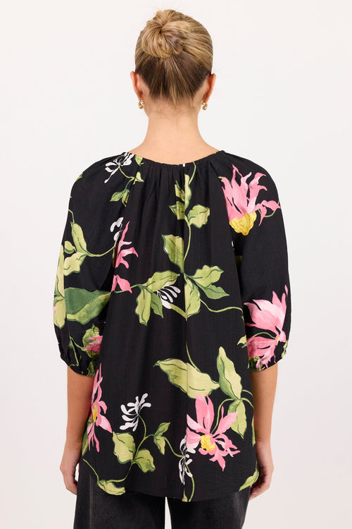 A woman viewed from behind wearing a black three-quarter sleeve top with a vibrant pink and green floral print, paired with black pants, against a plain white background.