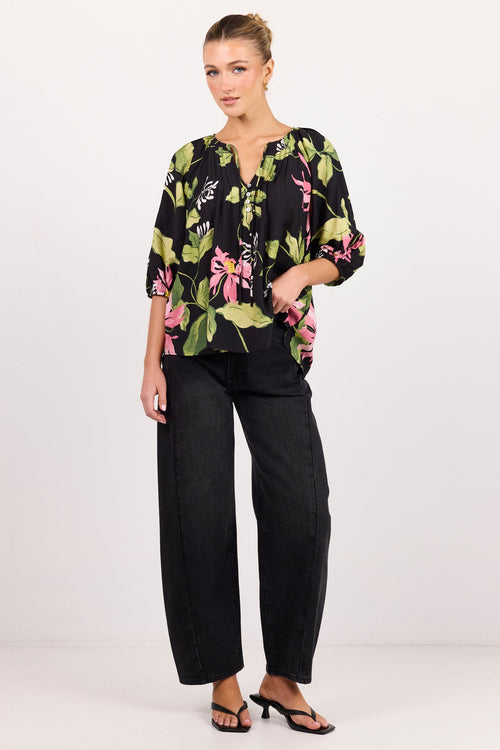 A woman modeling a black three-quarter sleeve blouse with a vibrant green, pink, and white tropical floral print, paired with black wide-leg trousers and black heeled sandals, against a plain white studio background.