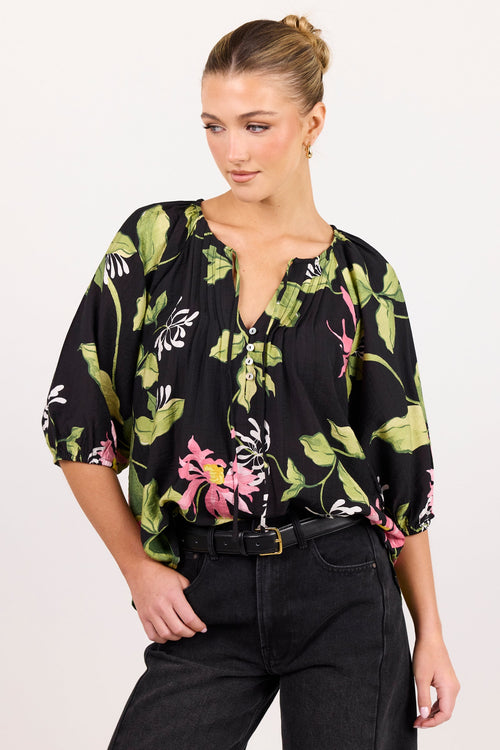 A woman modeling a black short-sleeve blouse with a green and pink floral print, paired with black wide-leg jeans, against a plain white studio background.