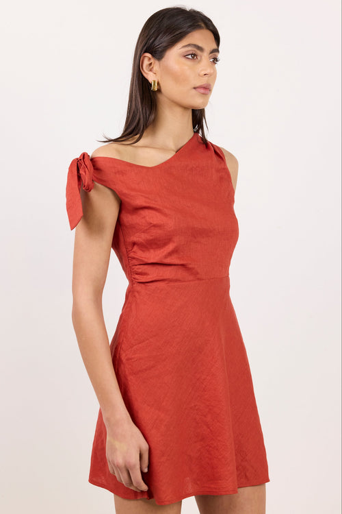 A woman modeling a rust-orange textured mini dress with an asymmetrical tie shoulder and a flared skirt, set against a plain light background.