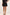 A model wearing a black high-waisted flared mini skirt with a matching black blouse and black sandals, shown from the waist down against a plain off-white studio background.