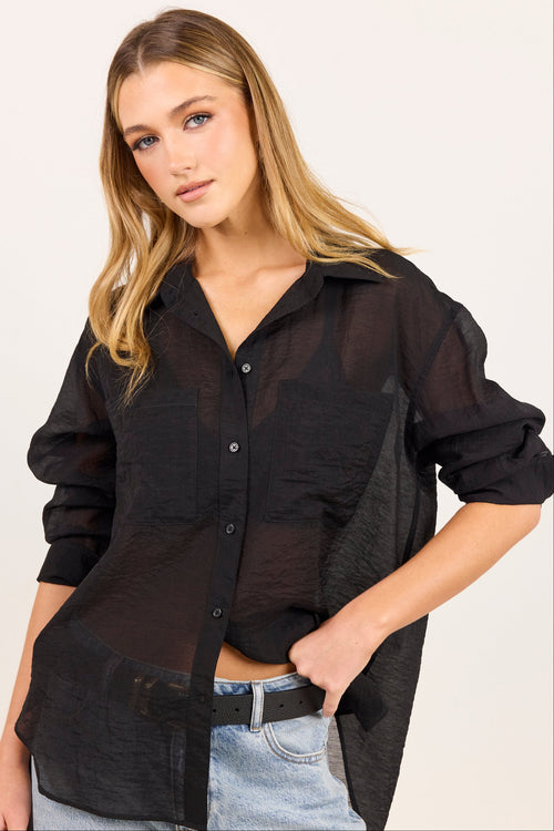 A woman wearing a black sheer button-up shirt with textured side panels, paired with light-wash jeans and a black belt, against a plain off-white background.