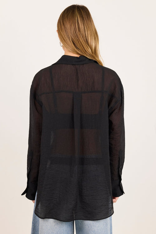 A woman viewed from behind wearing a sheer black long-sleeve button-up shirt over light blue jeans, set against a plain off-white studio background.