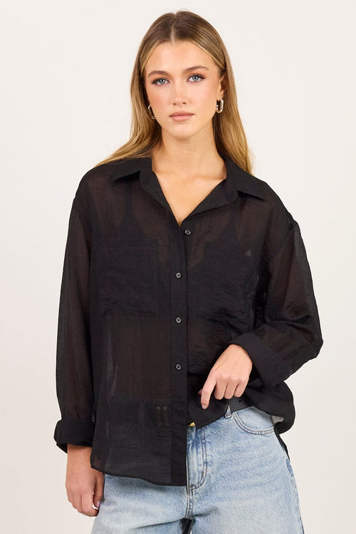 A woman modeling a black long-sleeve button-up shirt and light-wash blue denim culottes, paired with black strappy heels, against a plain off-white studio background.