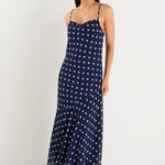 A model wears a navy blue bias-cut maxi dress with white polka dots, strappy sleeves, and a soft cowl neckline.
