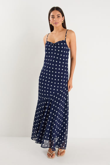 A model wears a navy blue bias-cut maxi dress with white polka dots, strappy sleeves, and a soft cowl neckline.