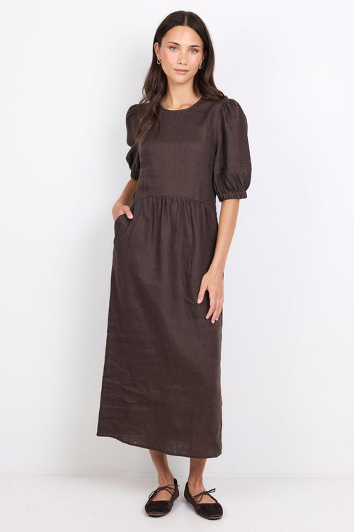 A woman modeling a dark brown midi dress with puffed short sleeves, a round neckline, and side pockets, standing against a plain white studio background.