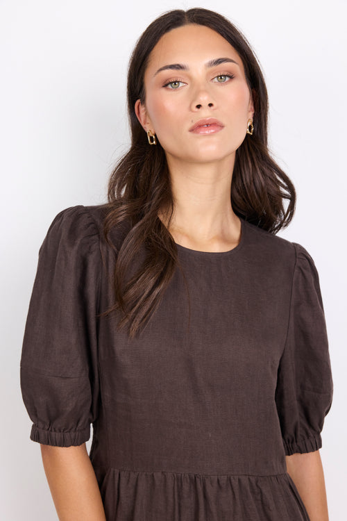 A woman wearing a dark brown puff-sleeve dress with a gathered waist, photographed against a clean white studio background.