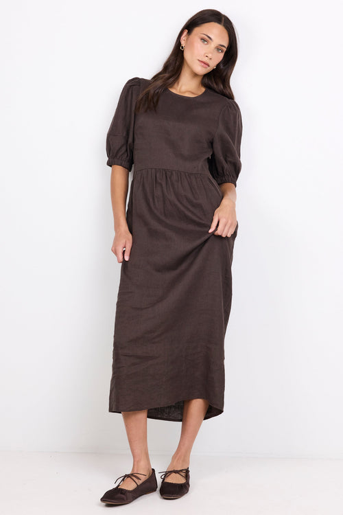 A woman modeling a dark brown midi dress with puff sleeves and a gathered waist, paired with dark brown flat shoes, against a plain white studio background.