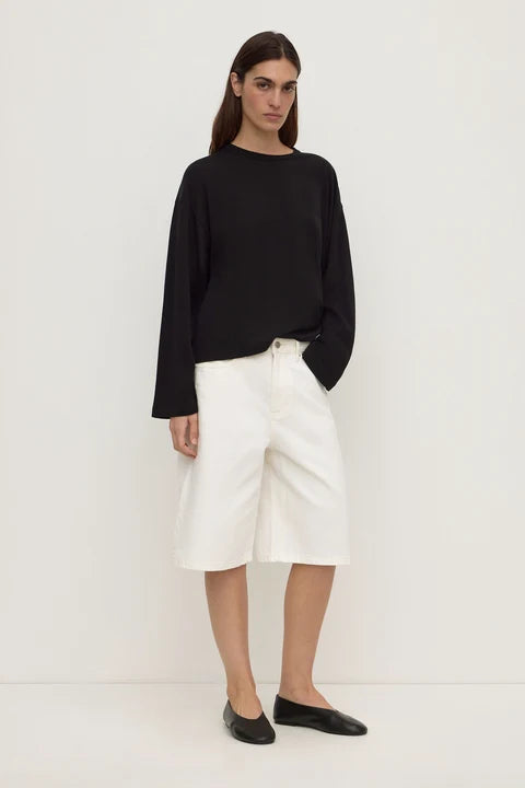A model stands against a plain white studio background wearing a black long-sleeve top, white cropped shorts, and black flats.