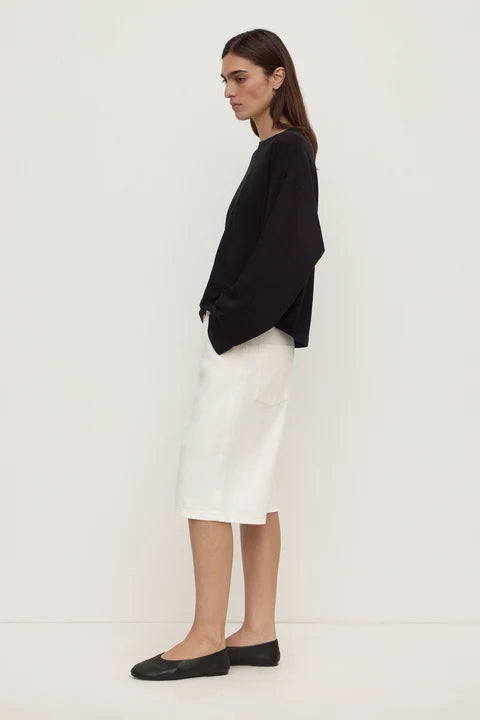 A woman stands in profile wearing a black long-sleeve top, a white knee-length skirt, and black flats against a plain off-white studio background.