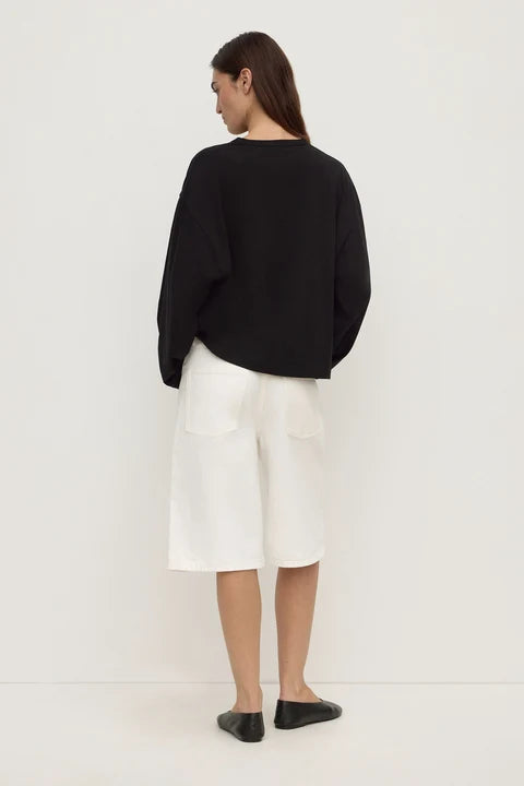 A woman shown from the back wearing a black long-sleeve cropped sweater, white wide-leg shorts, and black flats, standing against a plain white studio background.