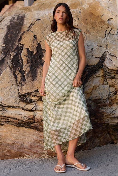 A woman wearing a light sage green and beige gingham midi dress with a sheer mesh overlay stands in front of a large rock formation outdoors.