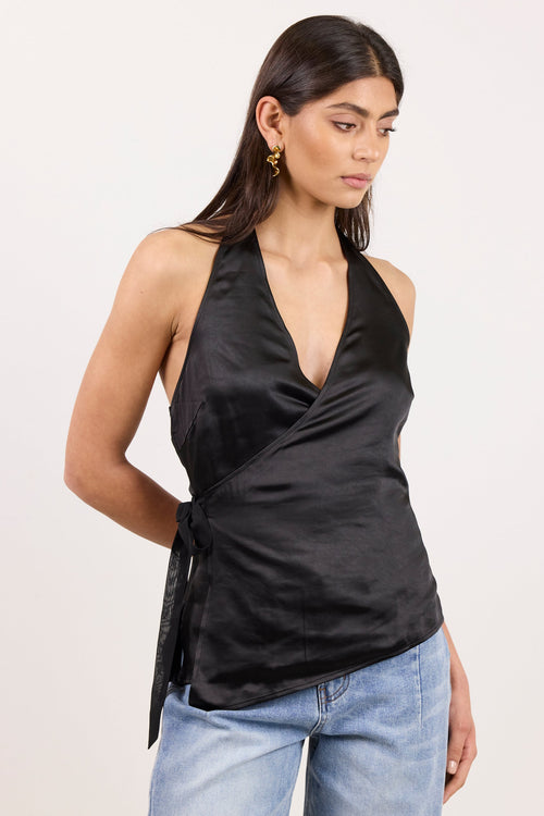 A woman modeling a black satin halter wrap top with a side tie, paired with light blue jeans, against a plain off-white studio background.