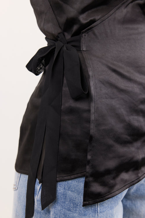 Close-up of a black satin wrap-style top tied at the waist with long ribbon tails, worn over light blue jeans against a plain light background.