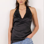 A woman modeling a black satin halter wrap top with light blue jeans, standing against a plain off-white background.