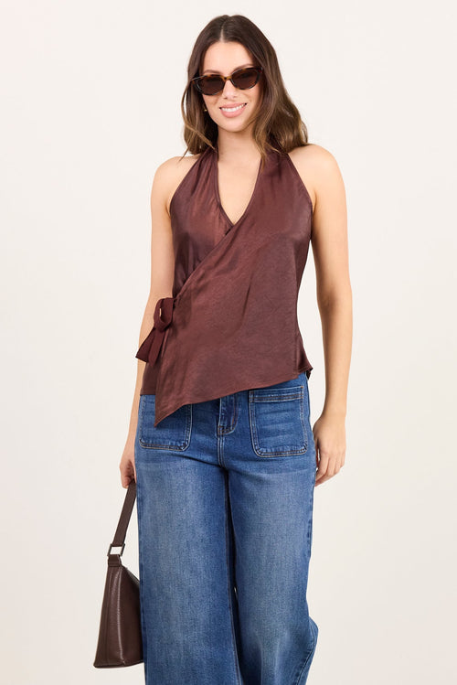 A woman modeling a sleeveless brown wrap-style top with side ties, paired with wide-leg blue jeans, black sandals, and a small brown handbag, standing against a plain light studio background.