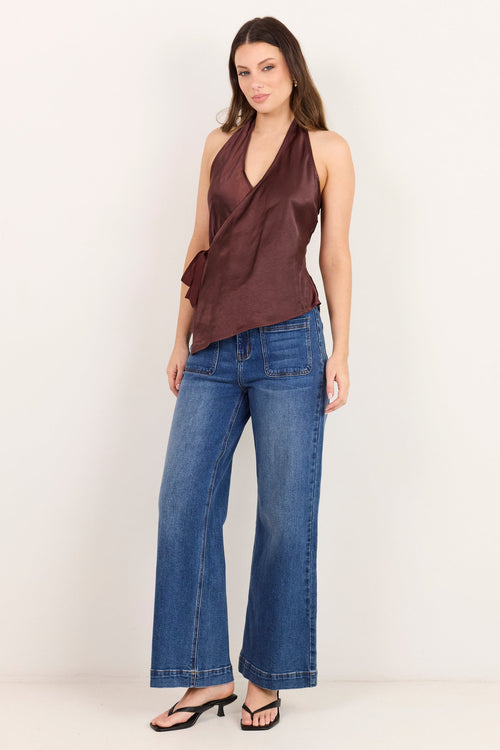 A woman modeling a dark brown halter wrap top and blue wide-leg jeans against a plain white studio background.