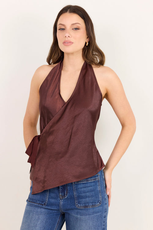 A woman modeling a sleeveless wrap-style halter top in a deep burgundy-brown color, paired with blue jeans, against a plain off-white studio background.