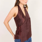 A woman wearing a sleeveless halter-neck wrap top in a deep brownish-rose color, paired with blue jeans, standing against a plain off-white background.